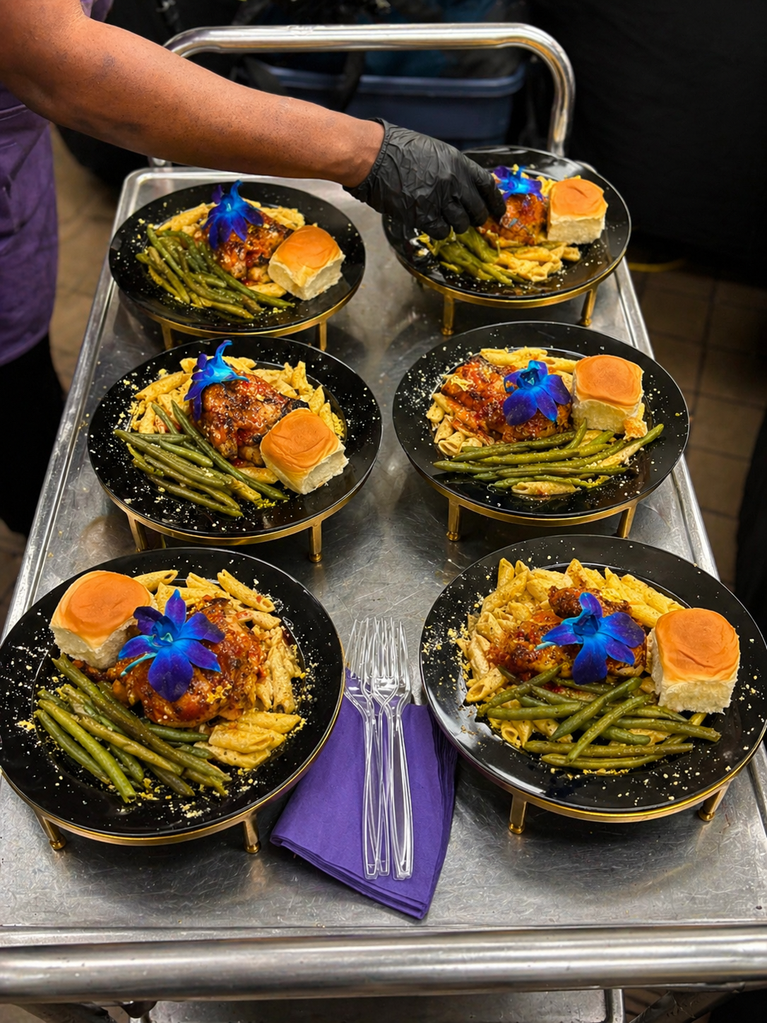 Plated arena catering — jerk chicken, pasta, green beans with blue orchid garnish on black and gold plates